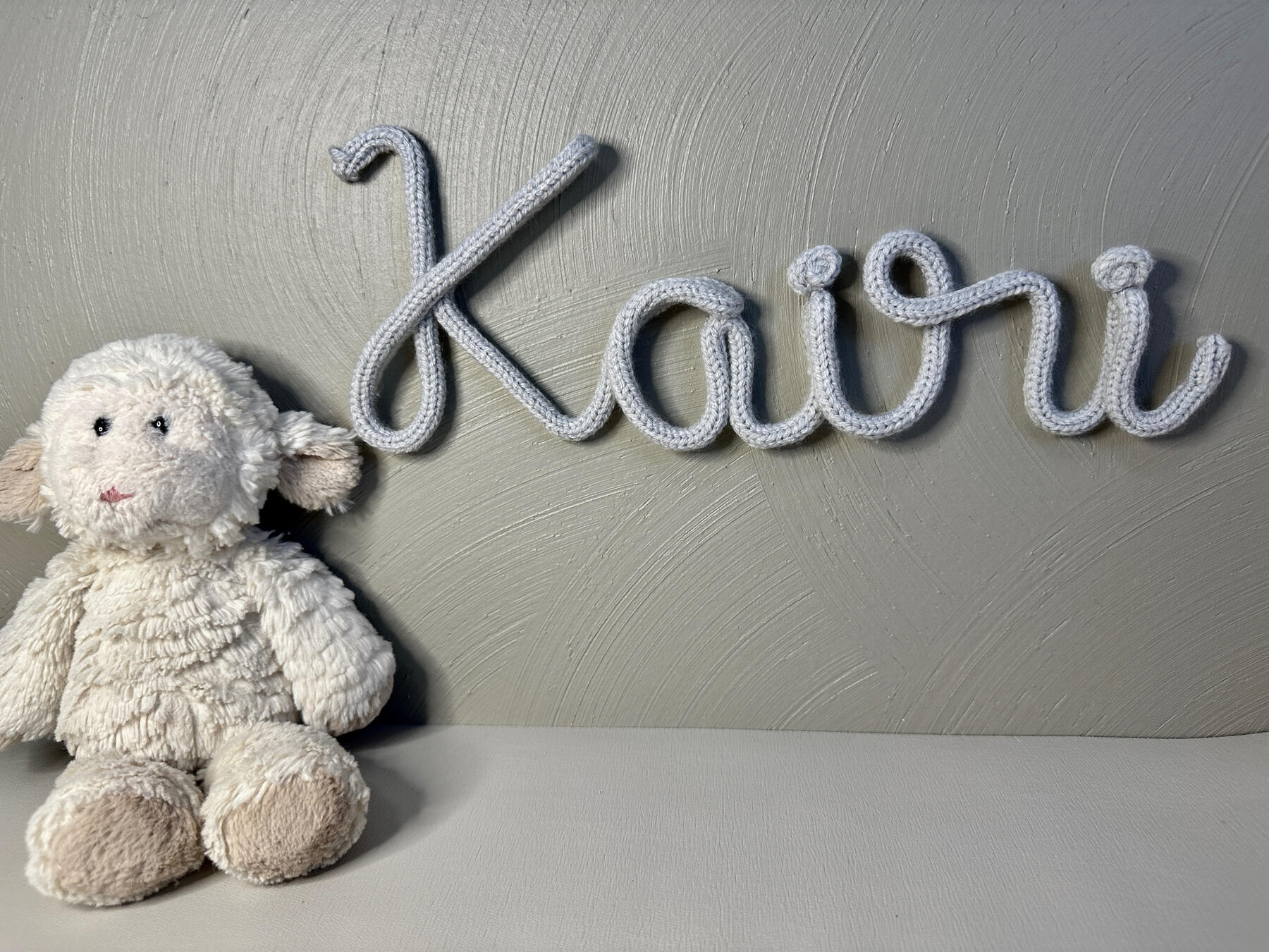 Kairi with gray yarn hung on the wall with a lamb stuffed animal sitting on a shelf under the name